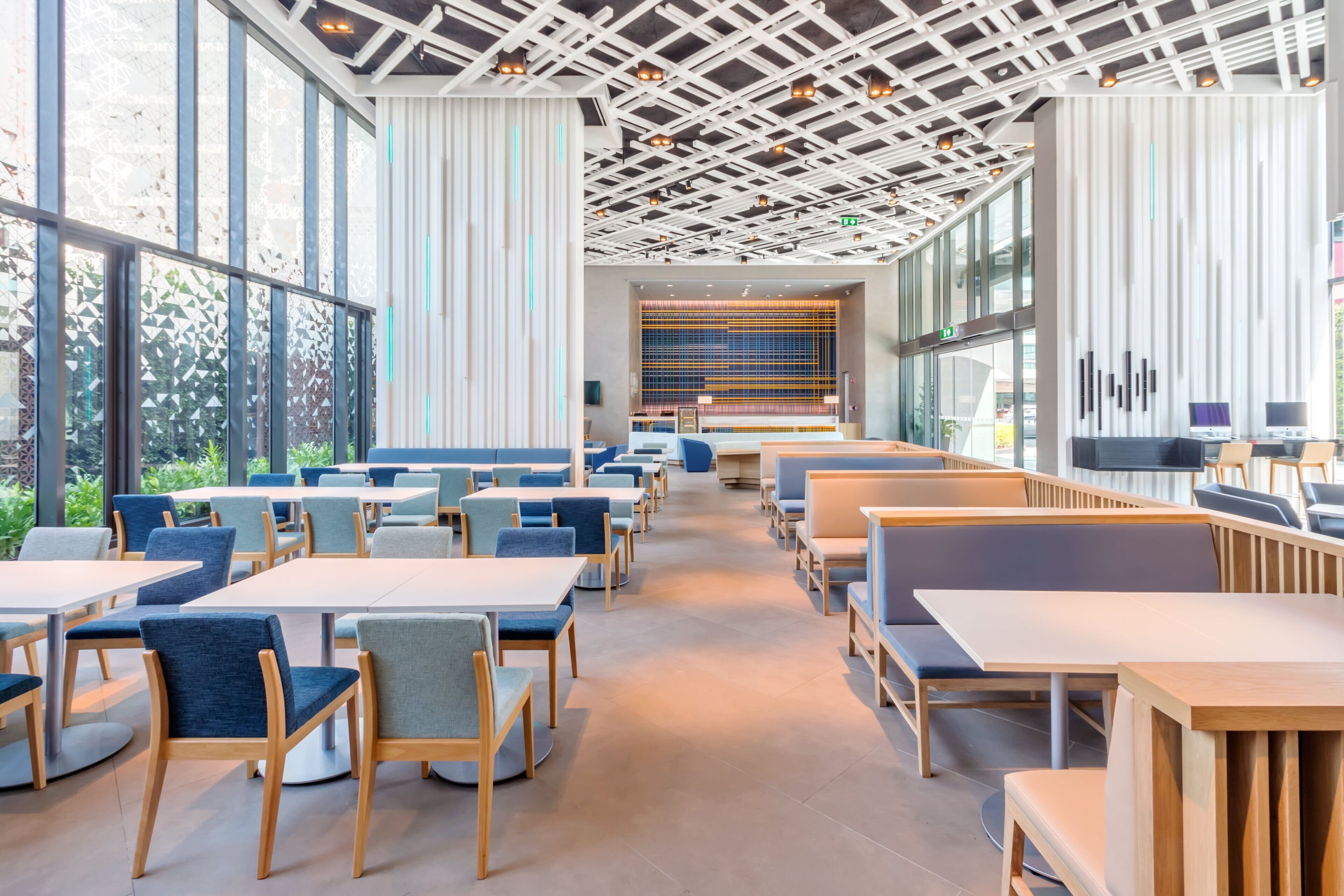 Un intérieur moderne avec des tables et des chaises disposées pour les repas, de grandes fenêtres laissant entrer la lumière naturelle et un plafond orné de poutres apparentes et de luminaires.