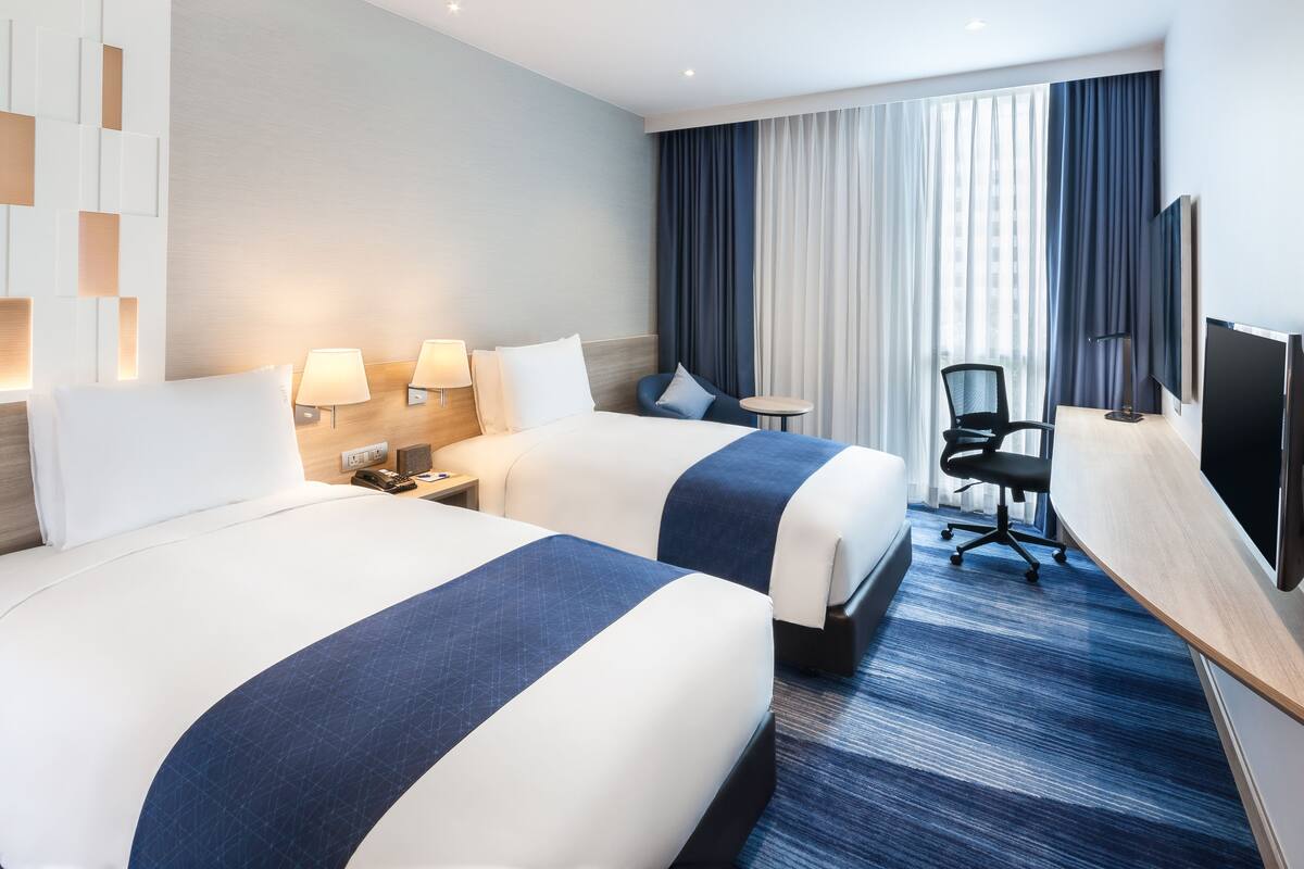 A hotel room with two beds, a desk, a chair, a TV, and a large window with curtains.