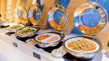 A buffet with various dishes, including pasta, salads, and meats, served in stainless steel bowls with lids.