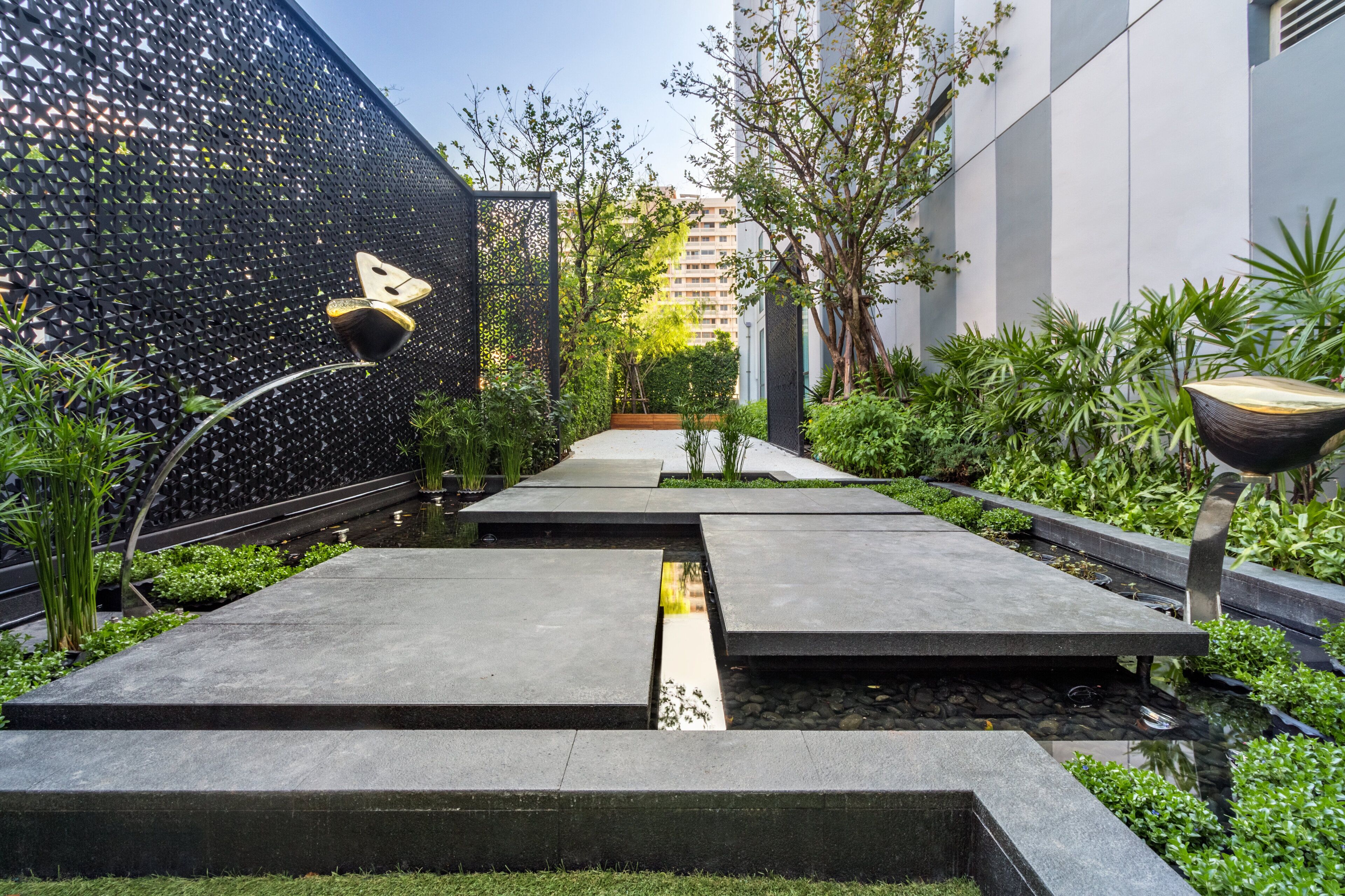 A modern outdoor pathway with geometric paving, surrounded by lush greenery and a decorative fence.