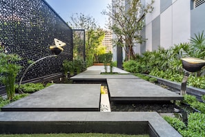 A modern outdoor pathway with geometric paving, surrounded by lush greenery and a decorative fence.