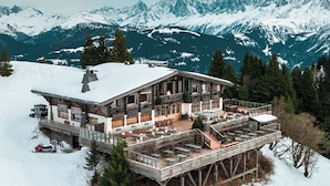 A wooden chalet with a balcony and outdoor seating area, surrounded by snow-covered mountains.