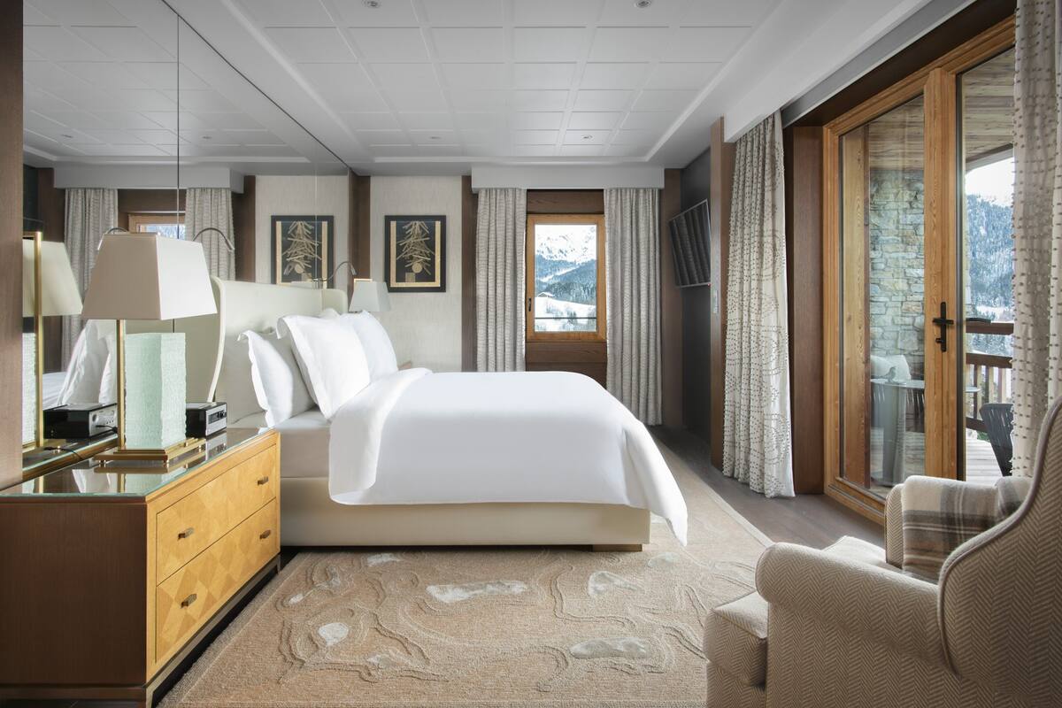 A modern hotel room with a large bed, a wooden dresser, a sitting area with a chair, and a view of mountains through a glass door.
