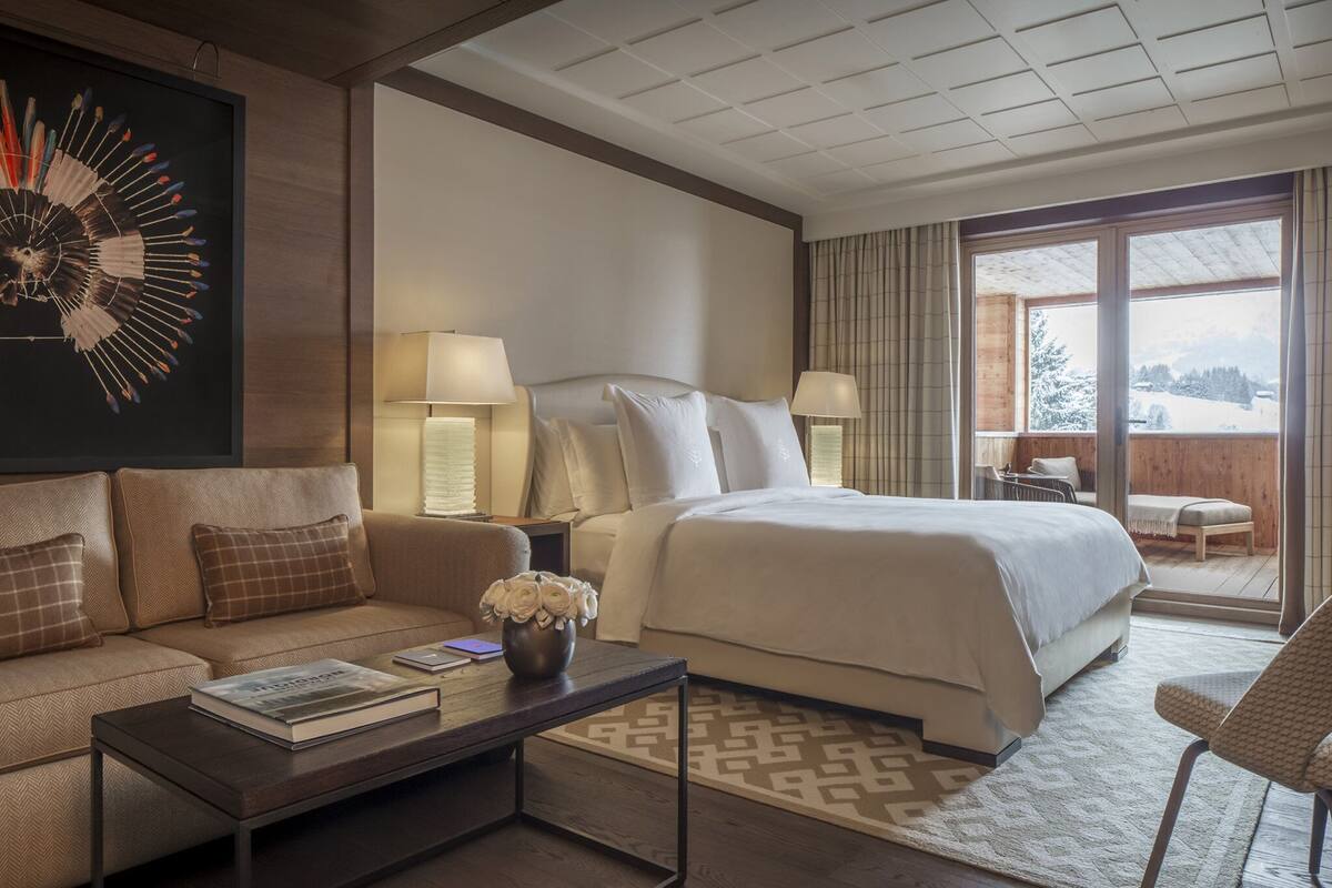 A modern hotel room with a large bed, a sofa, a coffee table with books, and a view of a snowy landscape through the window.