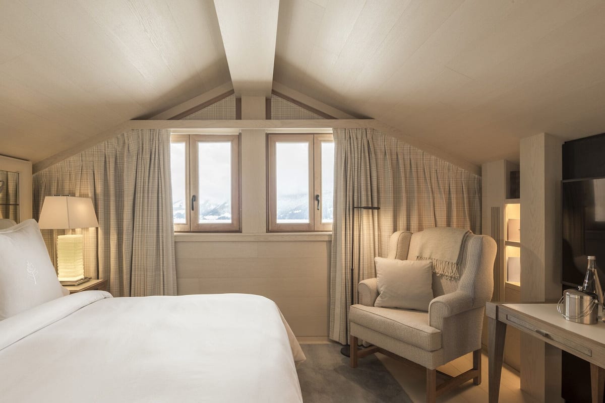 A hotel room with a large bed, a chair, a desk, and two windows with curtains.
