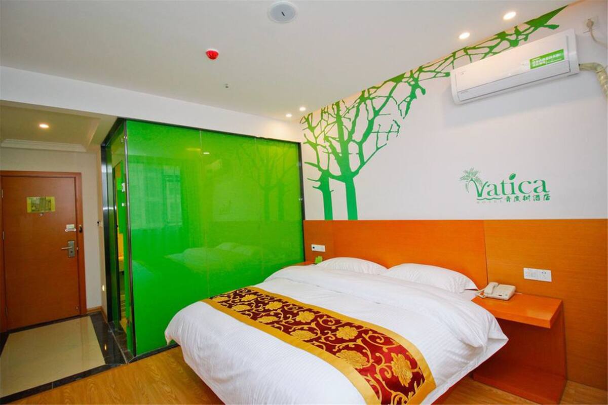A hotel room with a bed, a green partition, an orange headboard, a wall-mounted air conditioner, and a logo on the wall.