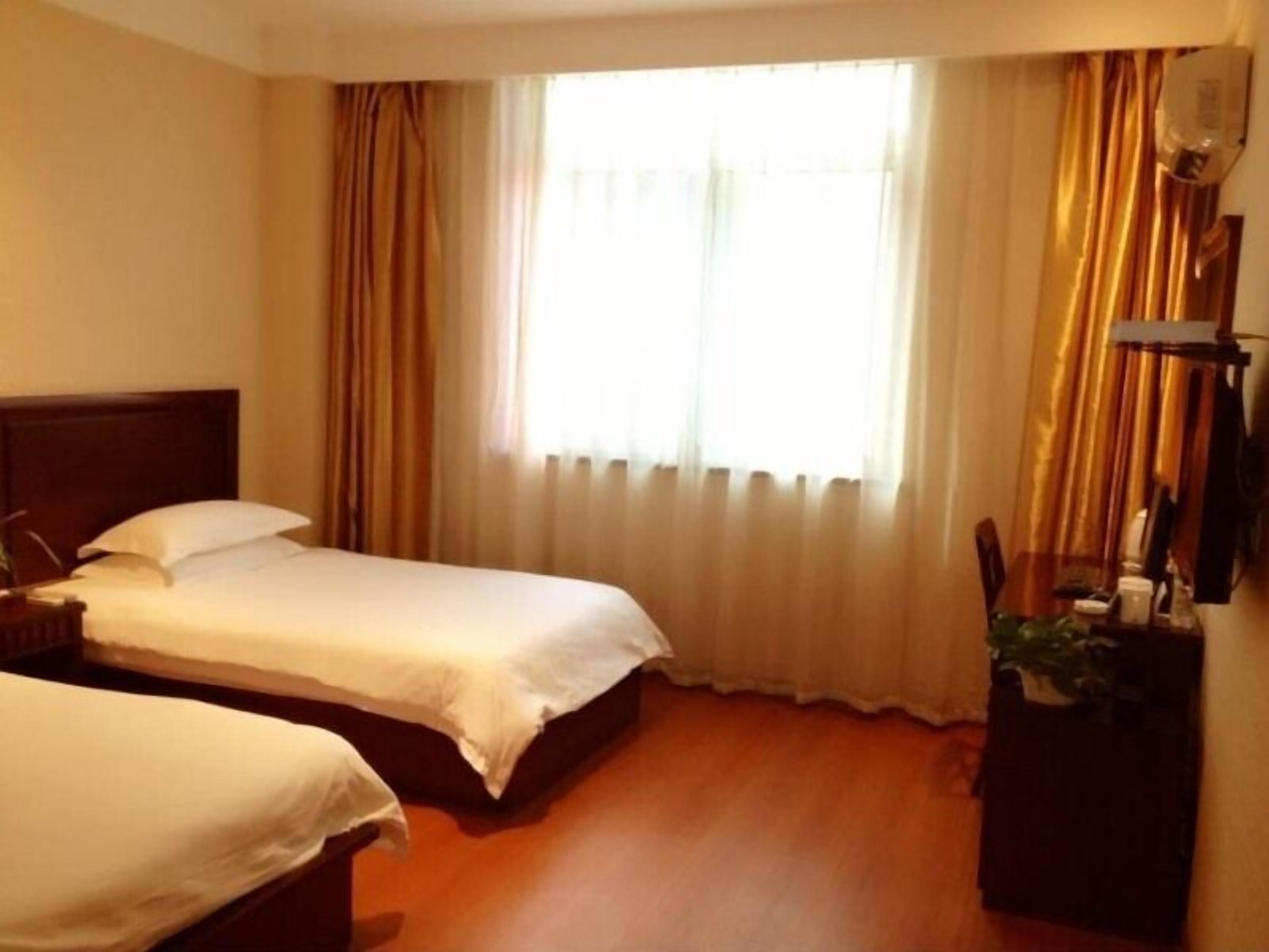 A hotel room with two single beds, a wooden floor, a large window with curtains, and a small desk with a plant.