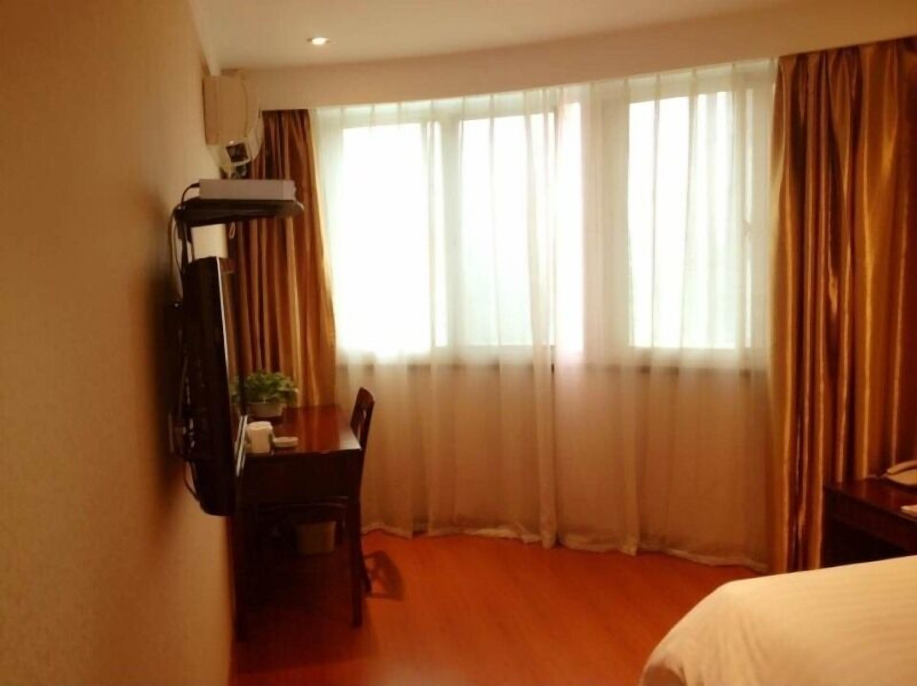 A hotel room with a bed, a desk with a chair, a TV mounted on the wall, and a window with curtains.