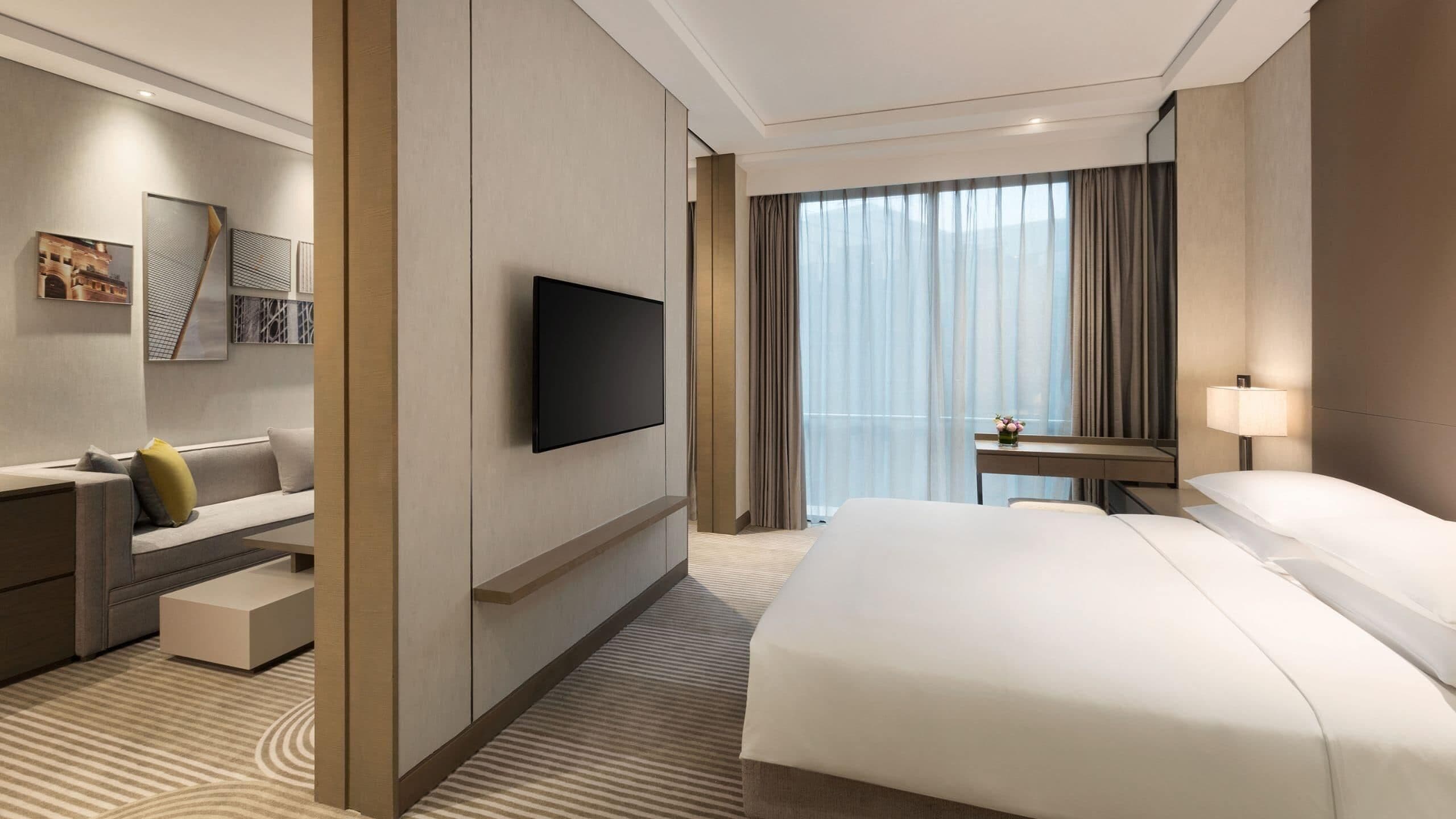 Una habitación de hotel moderna con una cama grande, un televisor de pantalla plana, un escritorio con una silla y vistas a la ciudad a través de amplias ventanas.