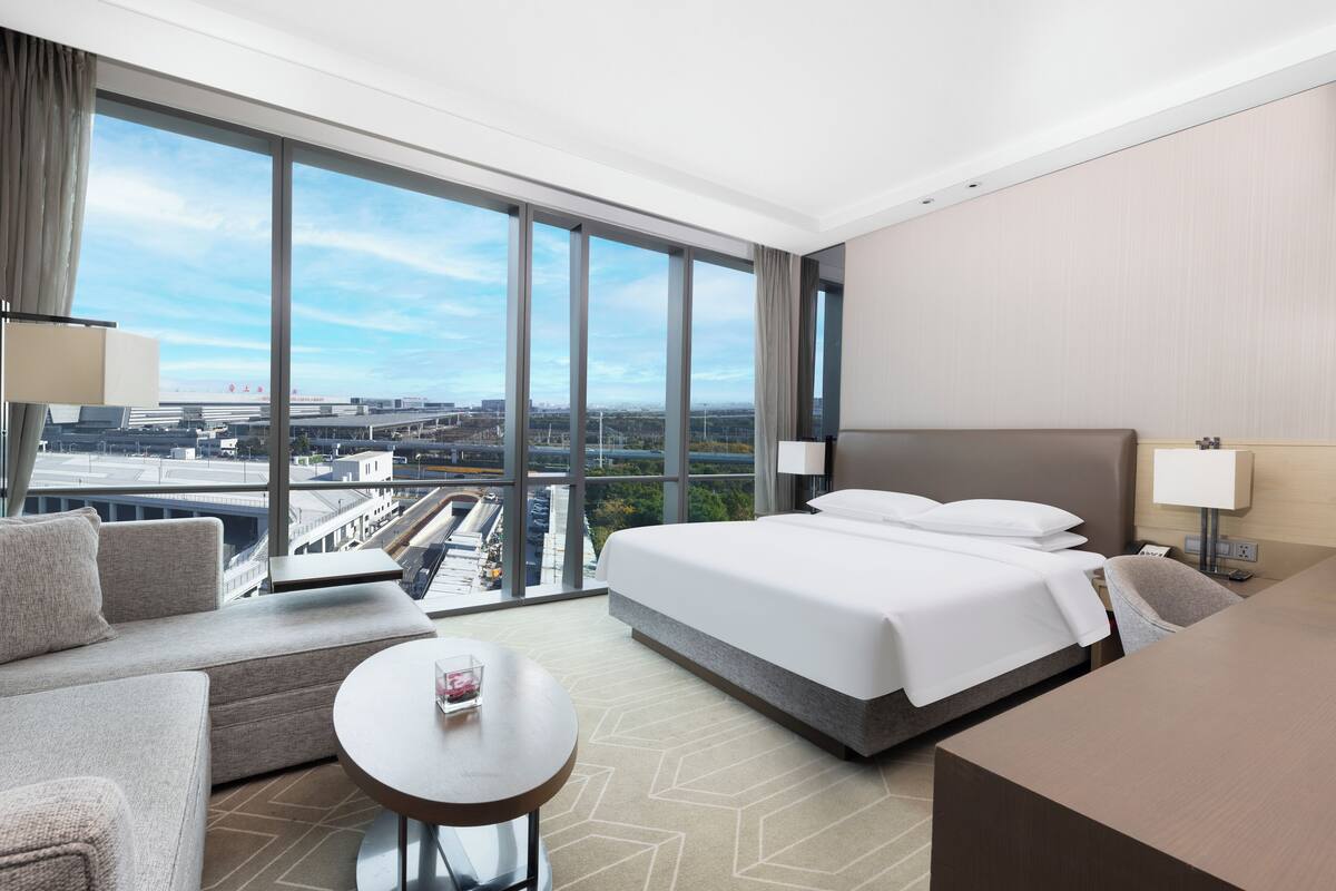 Une chambre d’hôtel avec un grand lit, un canapé, une petite table et une vue sur le paysage urbain, grâce à de grandes fenêtres.