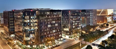 A modern multi-story building with a glass facade, surrounded by a landscaped area and a busy road with light trails indicating traffic.