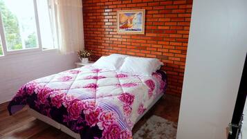 A bedroom with a brick wall, a bed with a floral bedspread, a nightstand with a vase, and a window with curtains.