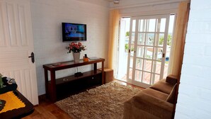 A living room with a television on a white brick wall, a wooden console table with a vase of flowers, a brown sofa, and a large window with a view of the outside. - Apartamento Vista do Centro Gramado 02 (Gramado)
