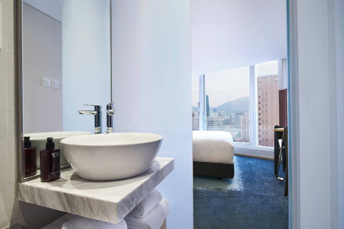 A modern bathroom with a large white sink, a mirror, and a view of a cityscape through large windows.