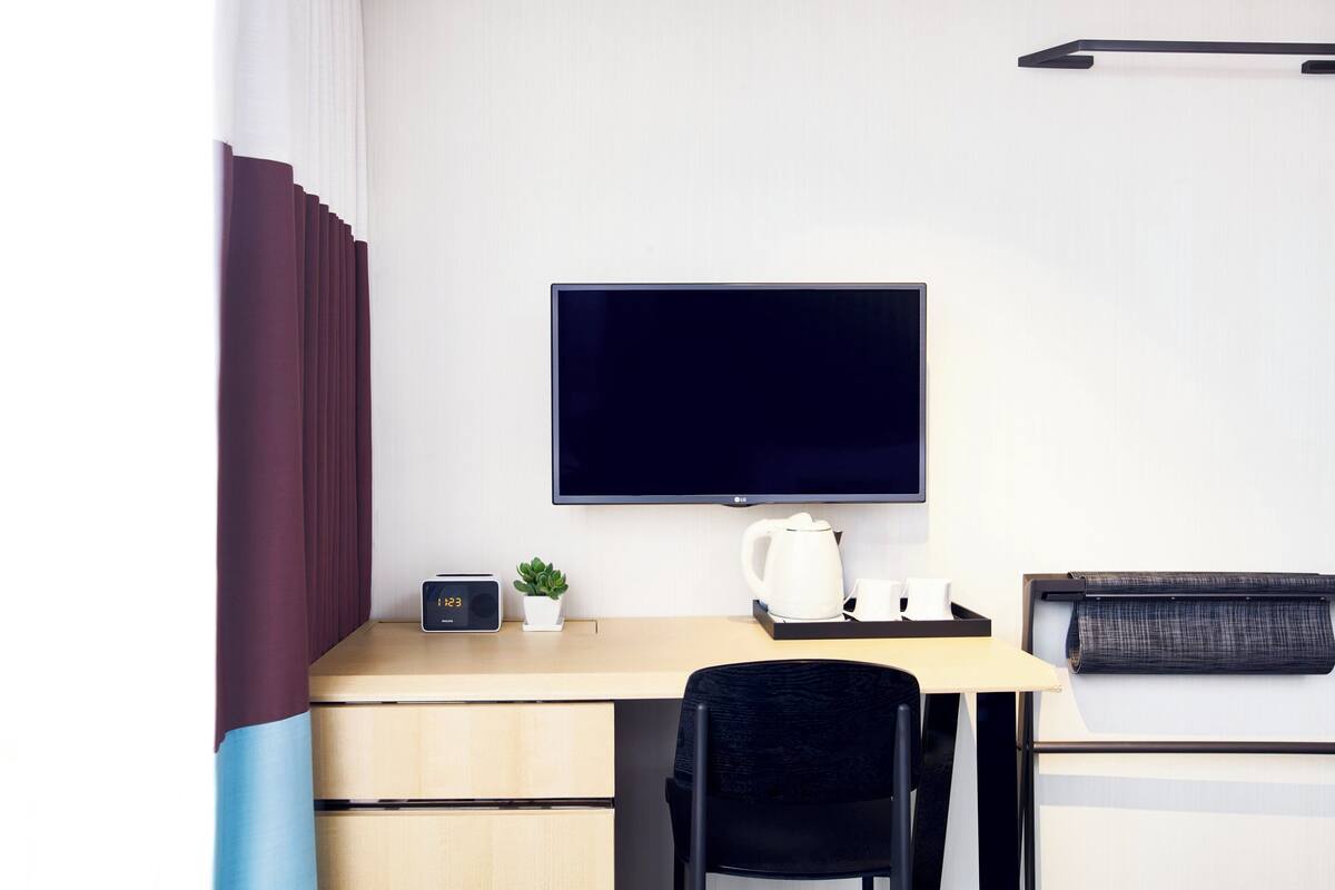 A modern hotel room with a desk, a flat-screen TV, a small plant, a kettle, and a neatly made bed.