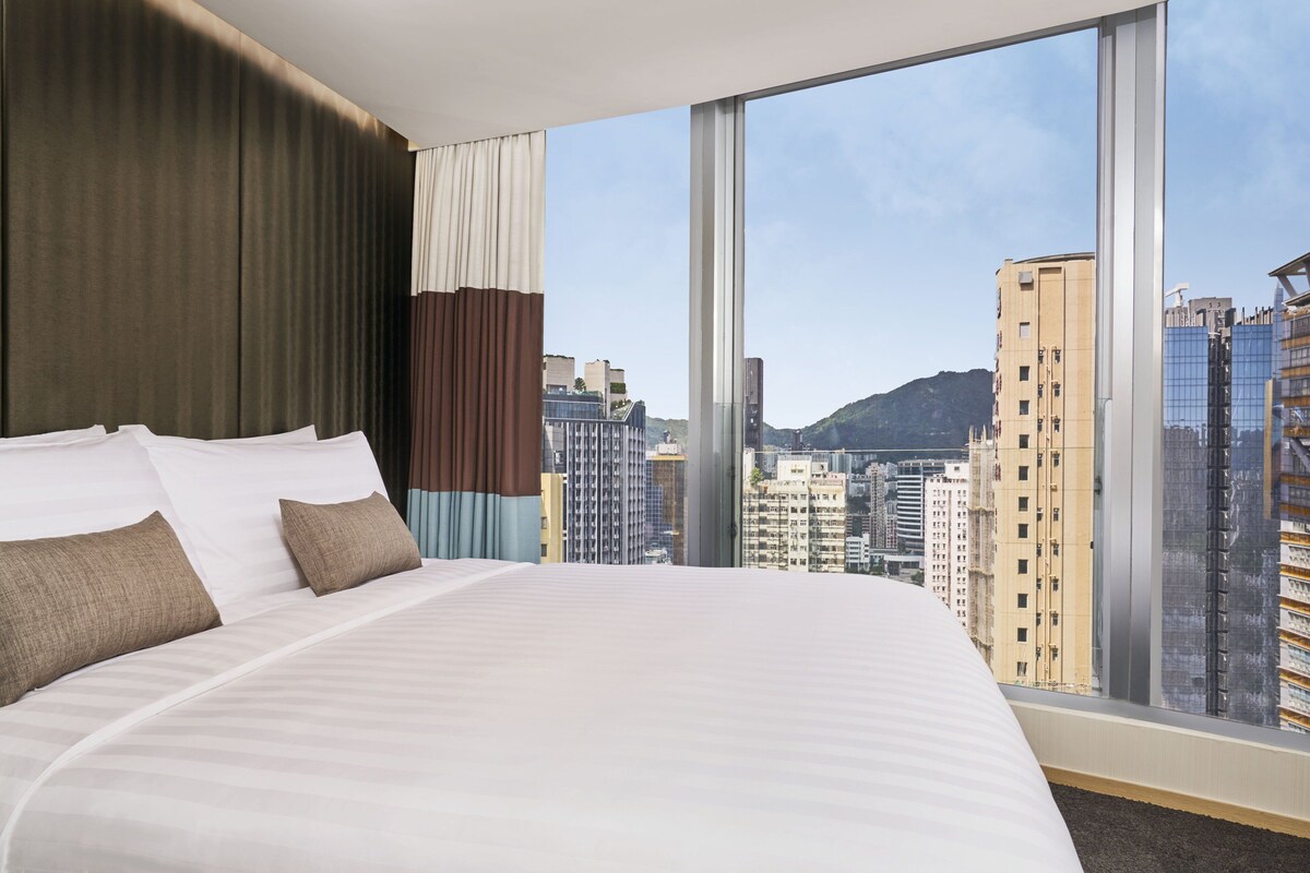 A hotel room with a large bed, a view of city buildings, and a window with curtains.