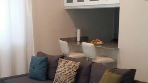 Een woonkamer met een donkergrijze bank, twee fauteuils, een eettafel met glazen blad en een keuken met witte kasten.