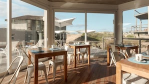 Un espacio exterior luminoso para comer, con mesas y sillas de madera, y amplias ventanas que ofrecen vistas a la playa y las dunas.
