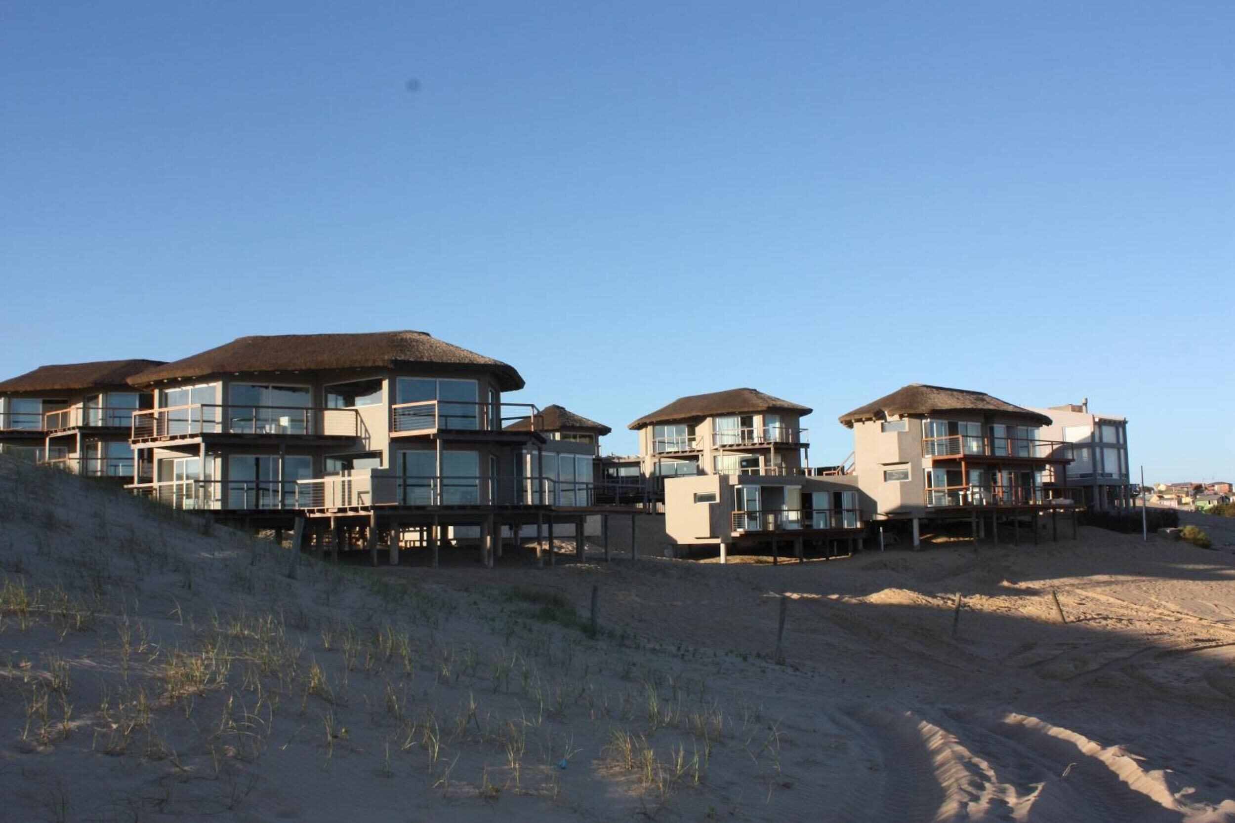 Propiedad frente a la playa con varios edificios de varios pisos, grandes ventanales y balcones, ubicada en un terreno arenoso con poca vegetación.