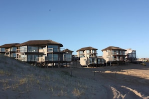 Propiedad frente a la playa con varios edificios de varios pisos, grandes ventanales y balcones, ubicada en un terreno arenoso con poca vegetación.