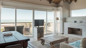 Amplia sala de estar con un gran ventanal corredero que ofrece vistas a la playa, una chimenea y un televisor sobre un mueble.