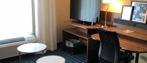A hotel room with a desk, two chairs, a TV, a lamp, and a window with curtains.