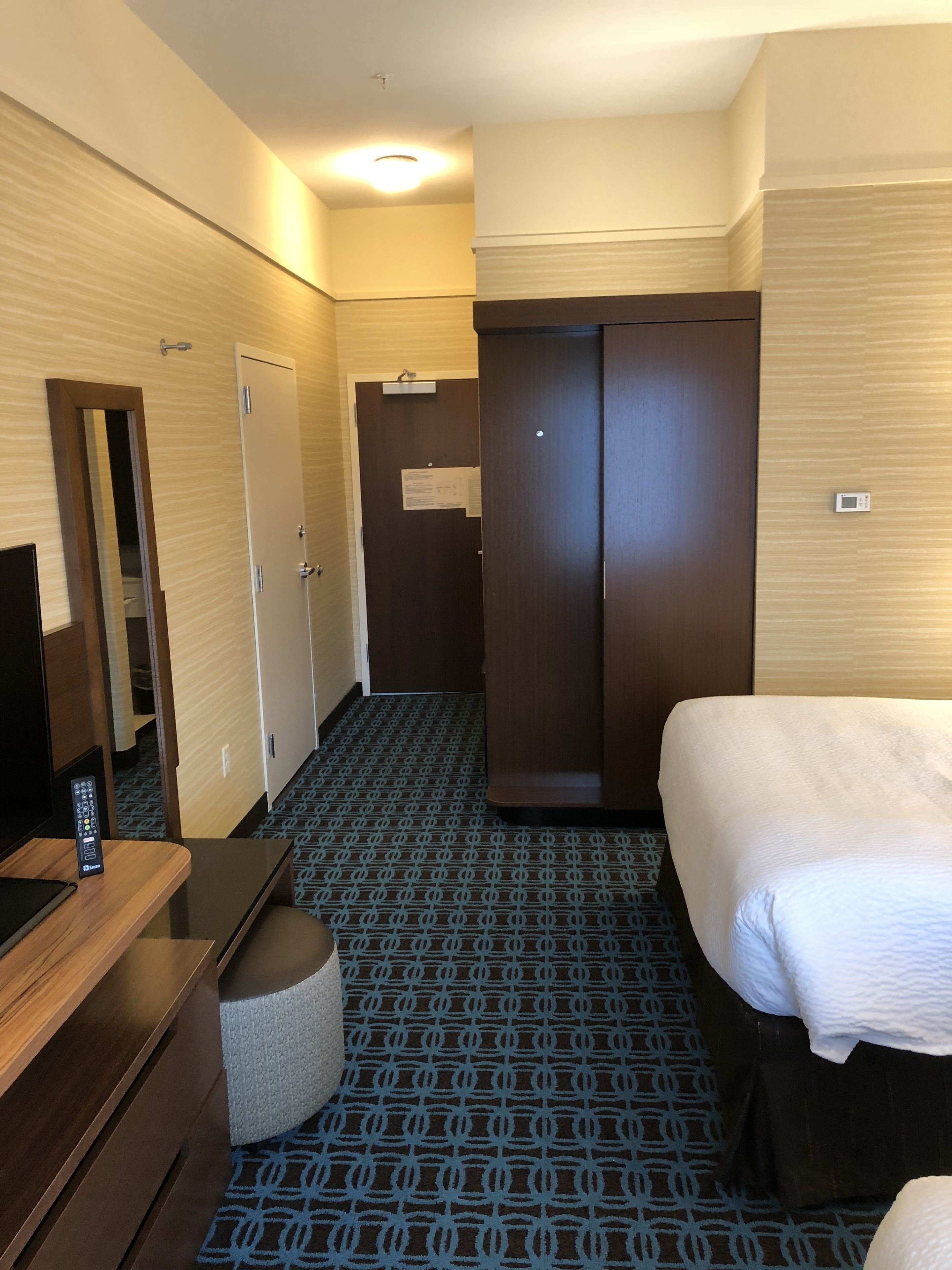 A hotel room with a bed, a desk with a remote control, a wardrobe, and a patterned carpet.