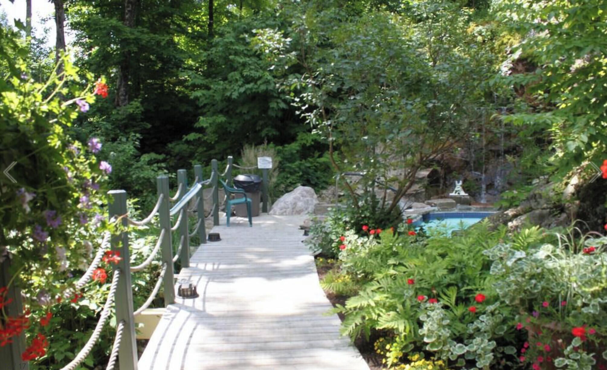 A wooden pathway leading to a pool area surrounded by lush greenery and flowering plants.