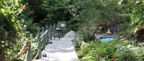 A wooden pathway leading to a pool area surrounded by lush greenery and flowering plants.