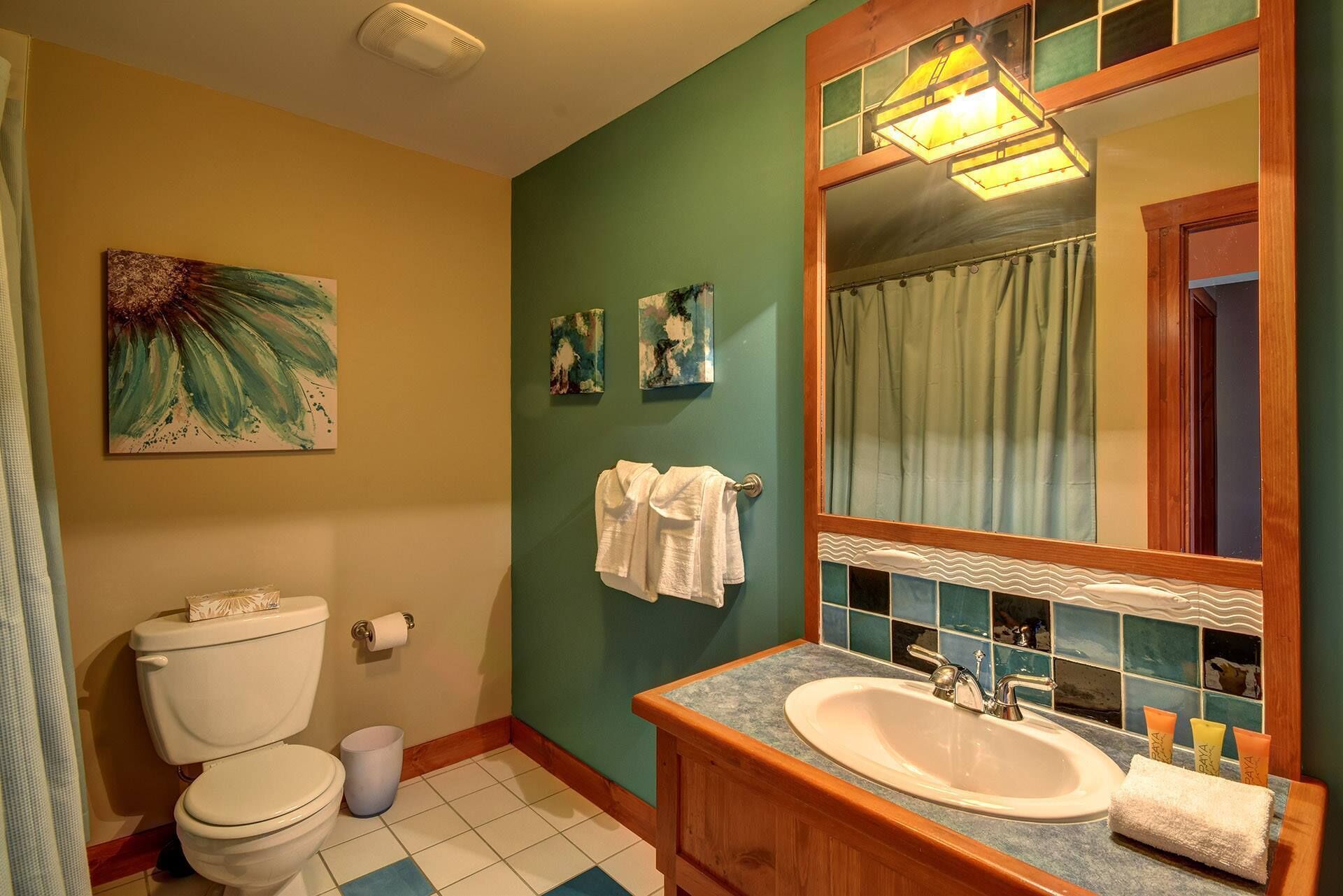 A bathroom with a toilet, sink, and mirror. There are towels hanging on the wall and a painting above the toilet.