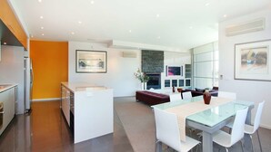 A modern kitchen with a central island, a dining area with a glass table, and a living space with a sofa and fireplace. - Penthouse St Kilda Style (St Kilda)