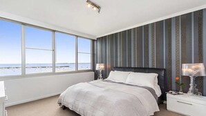 A bedroom with a large window offering a view of the beach, a bed with white bedding, and a nightstand with a lamp. - St Kilda Penthouse with Panaromic Bay and City View (St Kilda West)