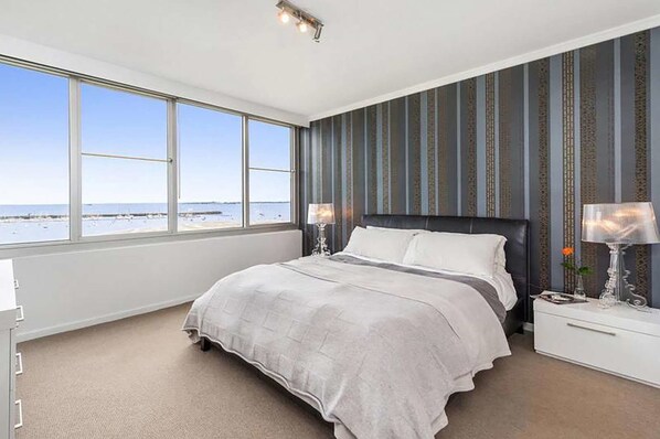 A bedroom with a large window offering a view of the beach, a bed with white bedding, and a nightstand with a lamp.