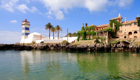 A coastal building with a lighthouse, palm trees, and a stone wall by the water.