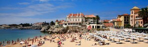 A beach with numerous umbrellas and sun loungers, a rocky shoreline, and buildings with red roofs in the background. - Cascais: apartment in the historic center, 3 minutes from the beach (Cascais)