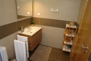 A modern bathroom with a wooden vanity, a white sink, and a mirror. There is a towel rack with a towel hanging, a small plant on the vanity, and a wooden shelf with baskets. - Cascais: apartment in the historic center, 3 minutes from the beach (Cascais)