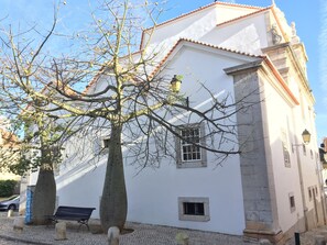 A white building with a tiled roof, a large tree in front, and a bench. - Cascais: apartment in the historic center, 3 minutes from the beach (Cascais)