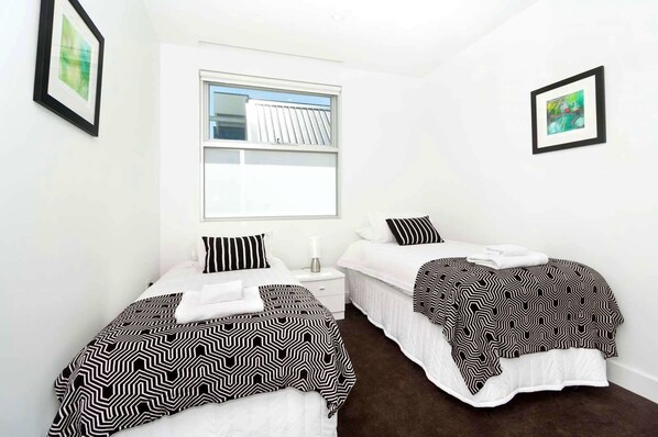 A hotel room with two beds, each with a black and white patterned bedspread and pillows. There is a window with blinds, a bedside table with a lamp, and a framed picture on the wall. - Broadway in Elwood on the Canal (Elwood)