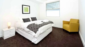 A bedroom with a bed, a bedside table, a chair, a window, and a framed picture on the wall. - Broadway in Elwood on the Canal (Elwood)