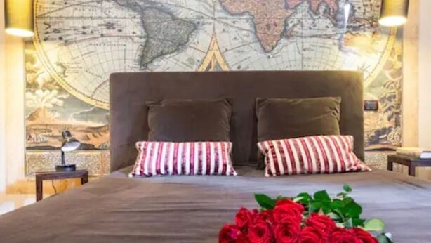 Une chambre avec une grande fresque représentant une carte du monde sur le mur, un lit avec une tête de lit marron, deux coussins à rayures et un bouquet de roses rouges posé sur le lit.