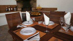 A dining table set with plates, glasses, and cutlery, with a stone fireplace and a fruit basket in the background. - Santa Eulalia Del Rio: chalet - IbizaHouse with character with pool and bbq place and magic seaview (santa eulalia del rio)