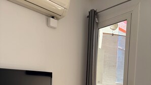 A room with a flat-screen TV mounted on the wall, a small white table with a vase and remote controls, and a sliding glass door leading to an outdoor patio area. - New apartment, 6 pers, free WiFi, Sagrada Familia (Barcelona)