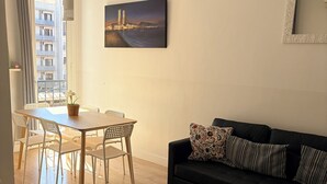 A modern living room with a dining area, a black sofa with patterned cushions, a cityscape painting, and a view of a building through a window. - New apartment, 6 pers, free WiFi, Sagrada Familia (Barcelona)