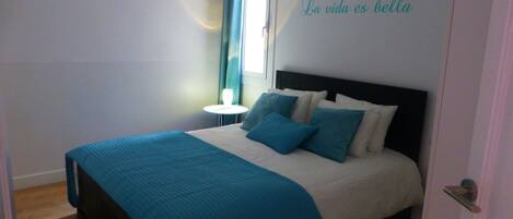 A neatly made bed with a blue bedspread and pillows, a small bedside table with a lamp, and a window with curtains.