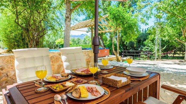 An outdoor dining area with a wooden table set for breakfast, including plates of eggs, bacon, and toast, along with glasses of orange juice.