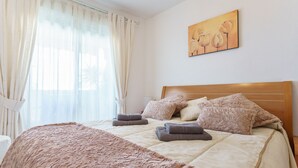 A neatly made bed with a beige quilt and a brown throw blanket, a wooden headboard, and a painting of flowers on the wall.
