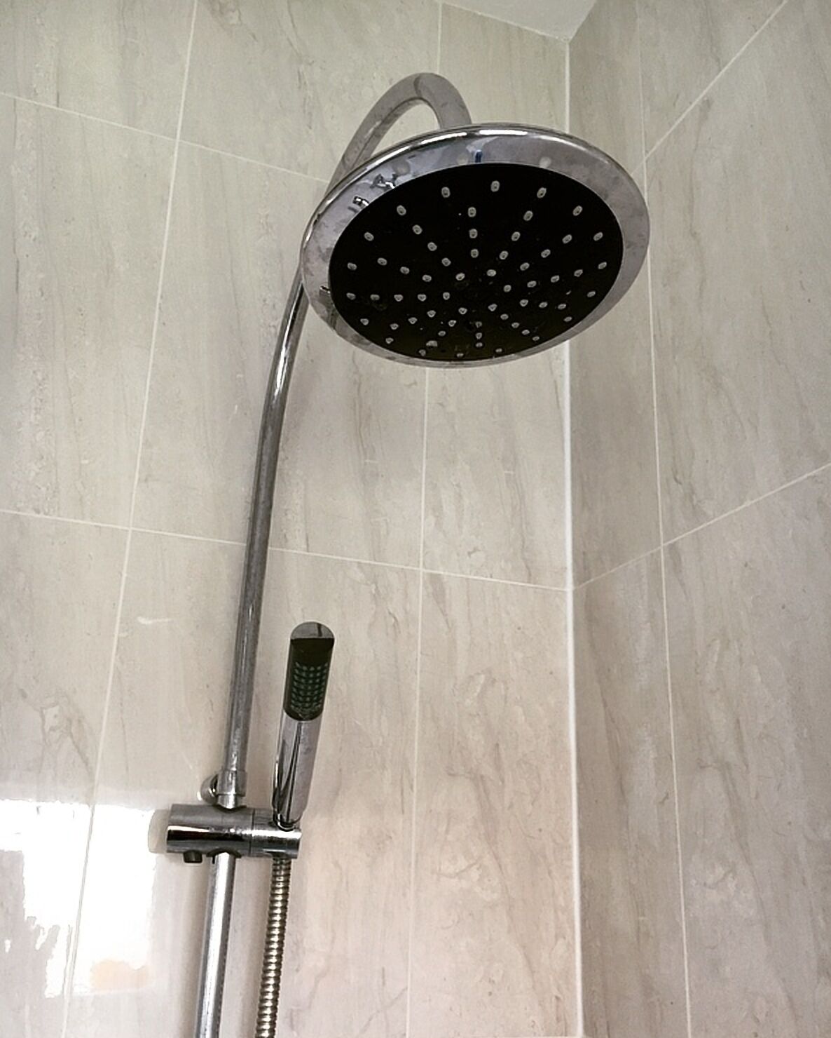 Un pommeau de douche doté d’un flexible, fixé sur un mur dont la surface est recouverte de carreaux.