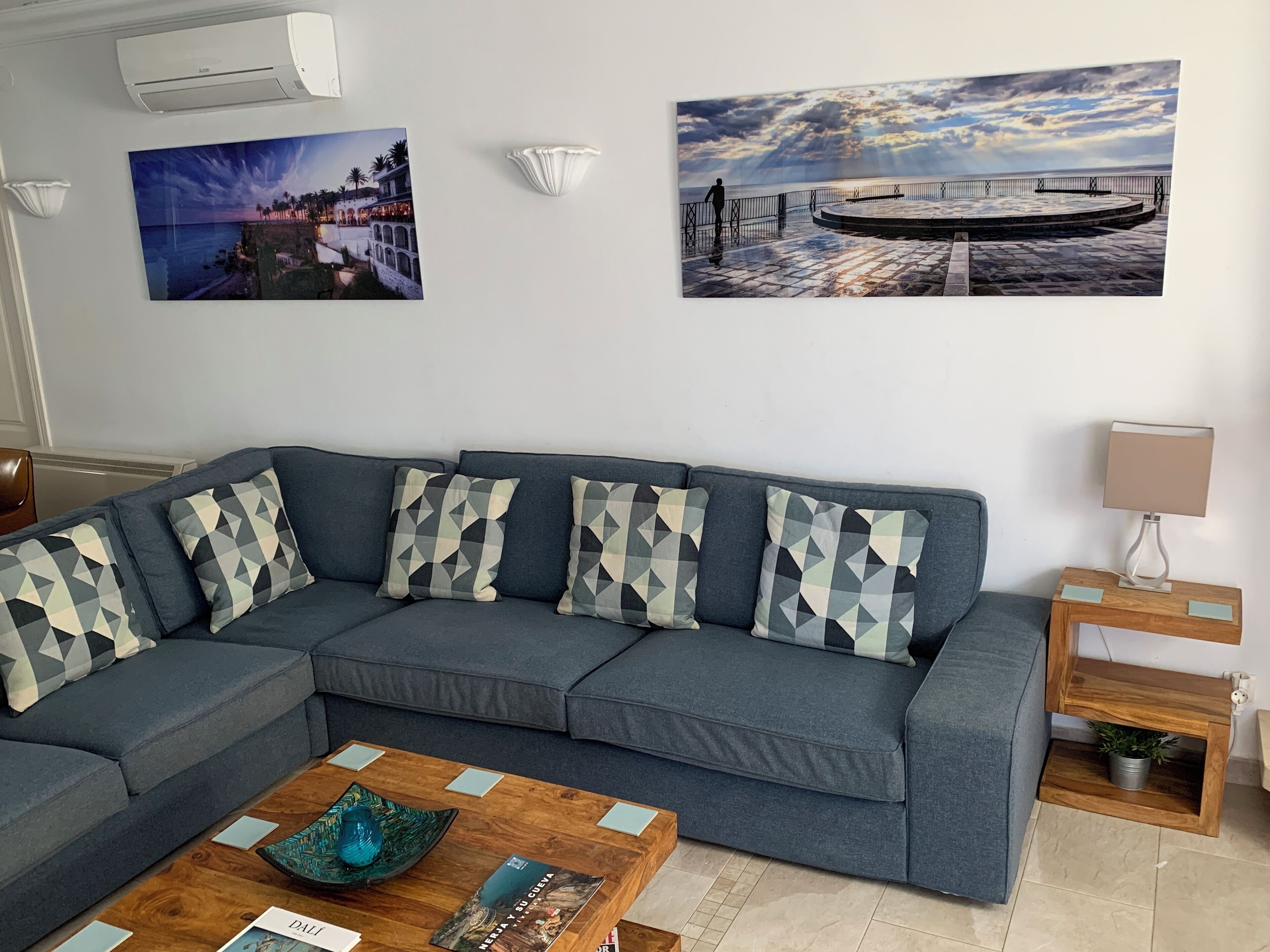 Una sala moderna con un sofá modular gris, una mesa de centro de madera y dos obras de arte colgadas en la pared que representan paisajes costeros.
