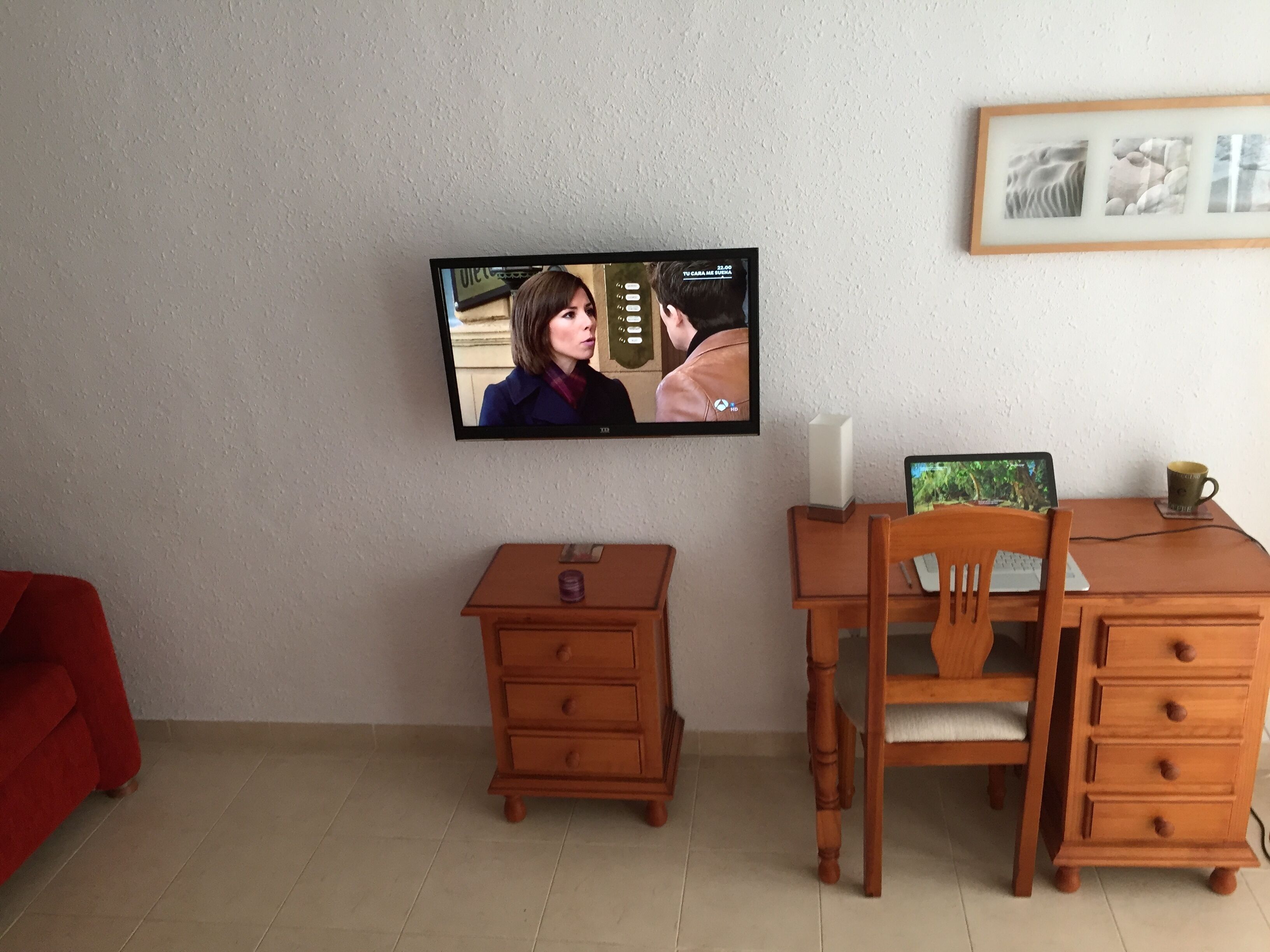 A room with a television mounted on the wall, a wooden desk with a chair, a small wooden nightstand, and a framed picture on the wall.