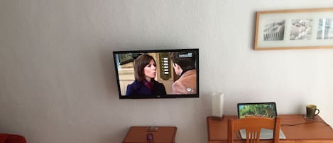 A room with a television mounted on the wall, a wooden desk with a chair, a small wooden nightstand, and a framed picture on the wall.